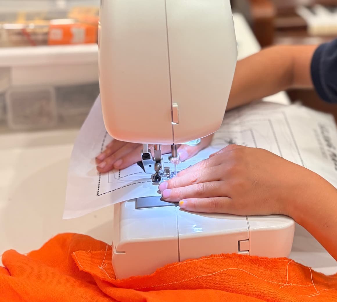 A young student guiding orange fabric under the needle of a sewing machine
