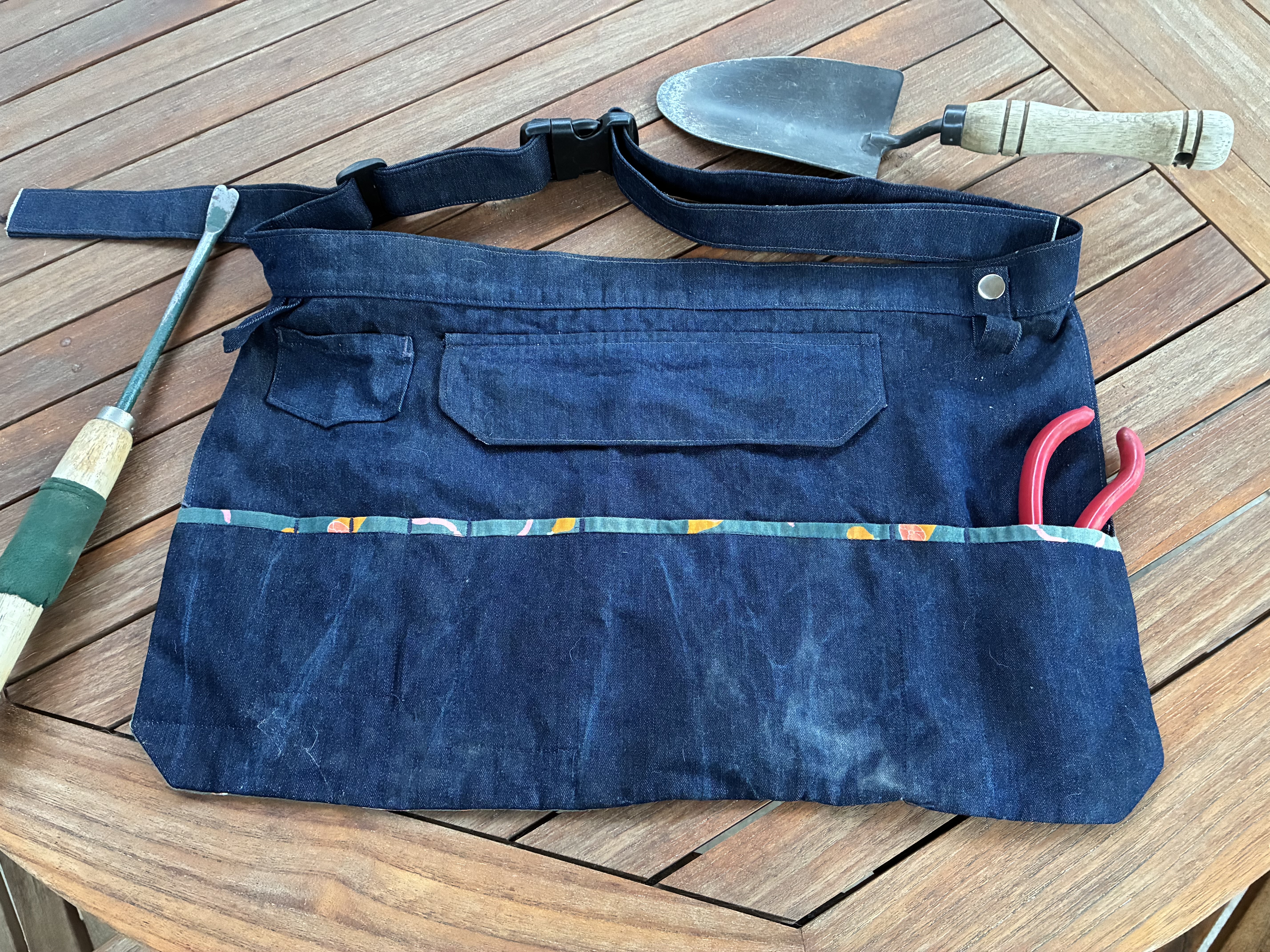 A denim waist-tie apron with gardening tools on a wooden table